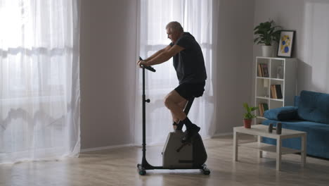El-Anciano-Está-Entrenando-En-Bicicleta-Estática-En-La-Sala-De-Estar,-Rehabilitación-Después-De-Una-Lesión,-Manteniendo-La-Salud-De-La-Respiración-Corporal-Y-El-Ejercicio-Cardiovascular.